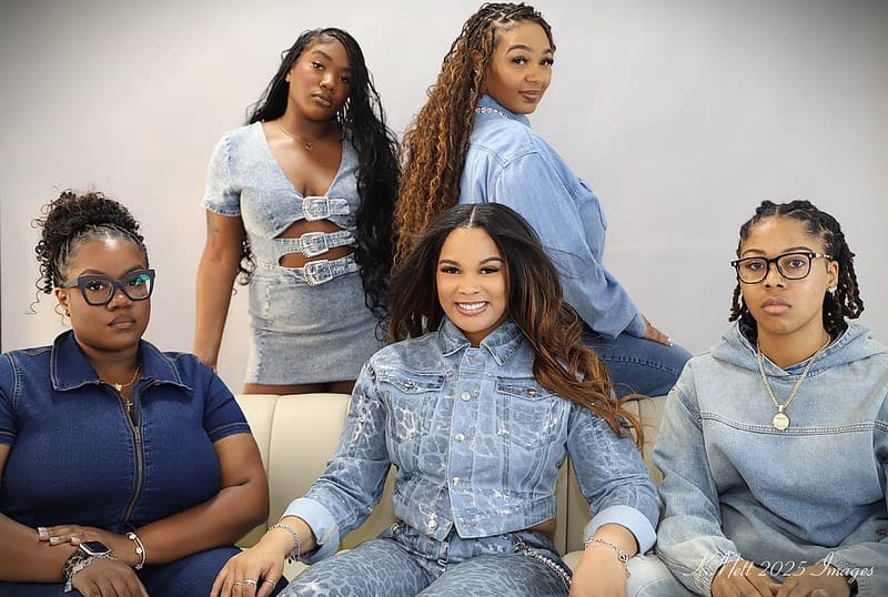 Kristian seated on a cream couch with four clients around her, everyone in denim. The styles range from long knotless braids to bohemian braids with curls left out and a braided updo.